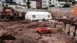 Eine Ausgrabungsfläche mit einem weißen Zelt und einem Bagger, im Hintergrund Wohnhäuser.jpg